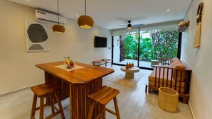 A modern dining area with a wooden table and bar stools, a comfortable seating area with cushions, and a large window offering a view of greenery. - Tú Refugio Exclusivo en Tulum Dk Jade 3 (Tulum)