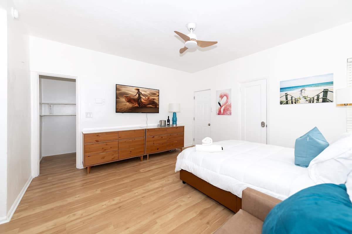 Une chambre à coucher comprenant un lit, une commode en bois, un ventilateur de plafond et une porte donnant sur une autre pièce.