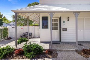 A white building with a covered porch, a door, and a small garden area with plants and gravel.