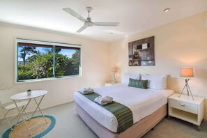 A hotel room with a bed, two bedside tables, a ceiling fan, a window with a view of trees, and a small table with a chair.