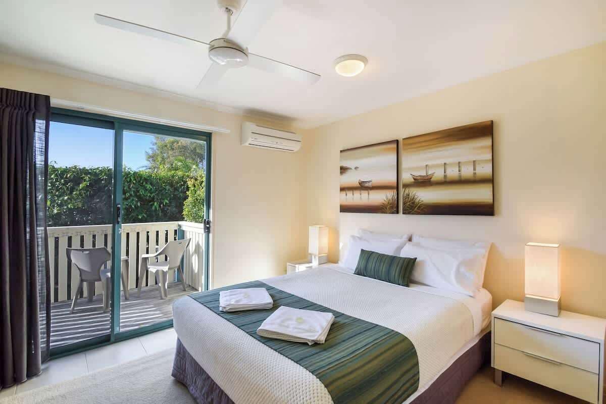 A hotel room with a bed, a nightstand, a lamp, a painting, and a balcony with chairs.