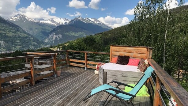 Terraza de madera con una cama y una silla de descanso, con vistas a un paisaje montañoso.