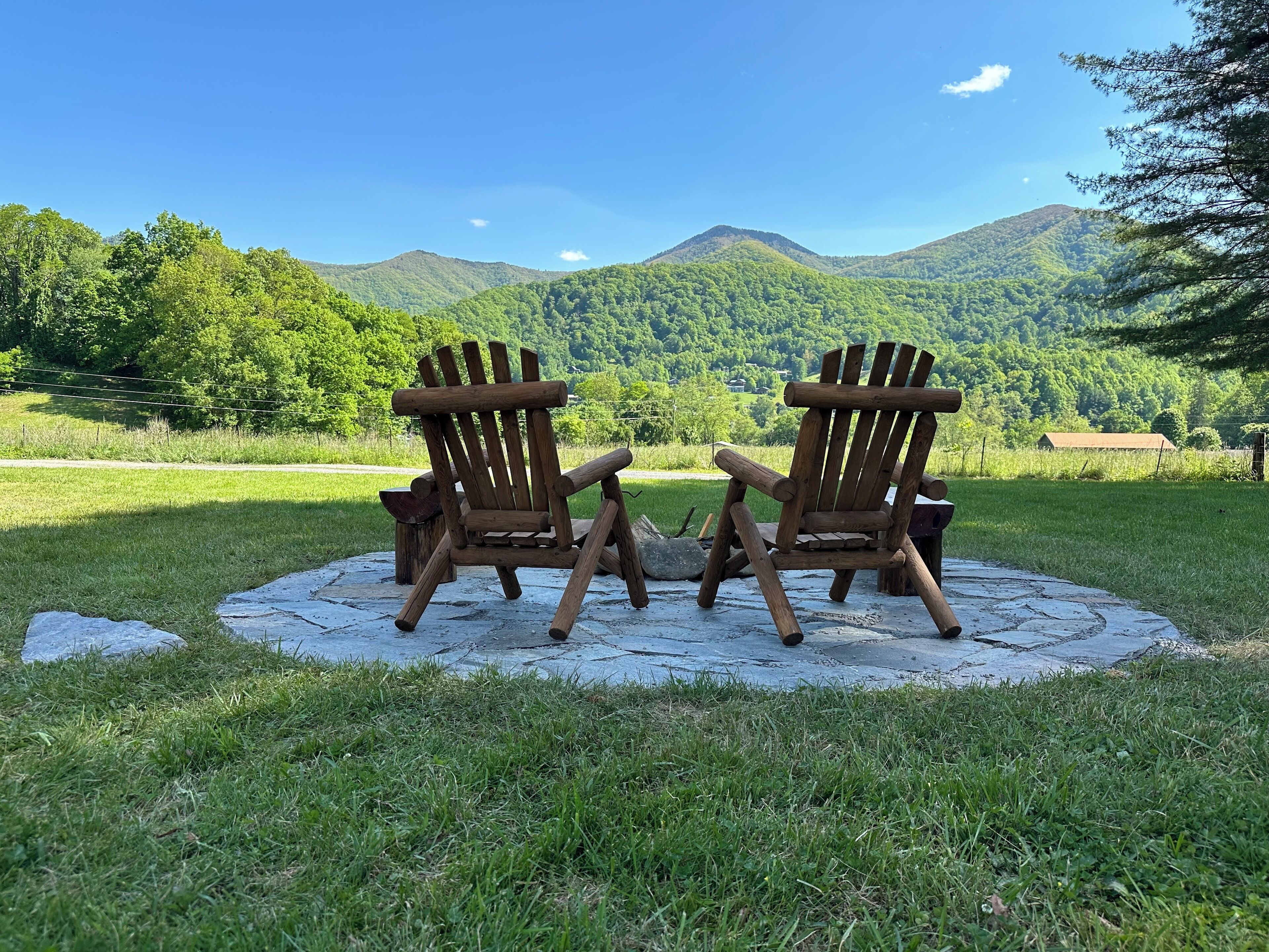 Two wooden Adirondack chairs facing each other on a concrete circle in a grassy field with a mountain backdrop.