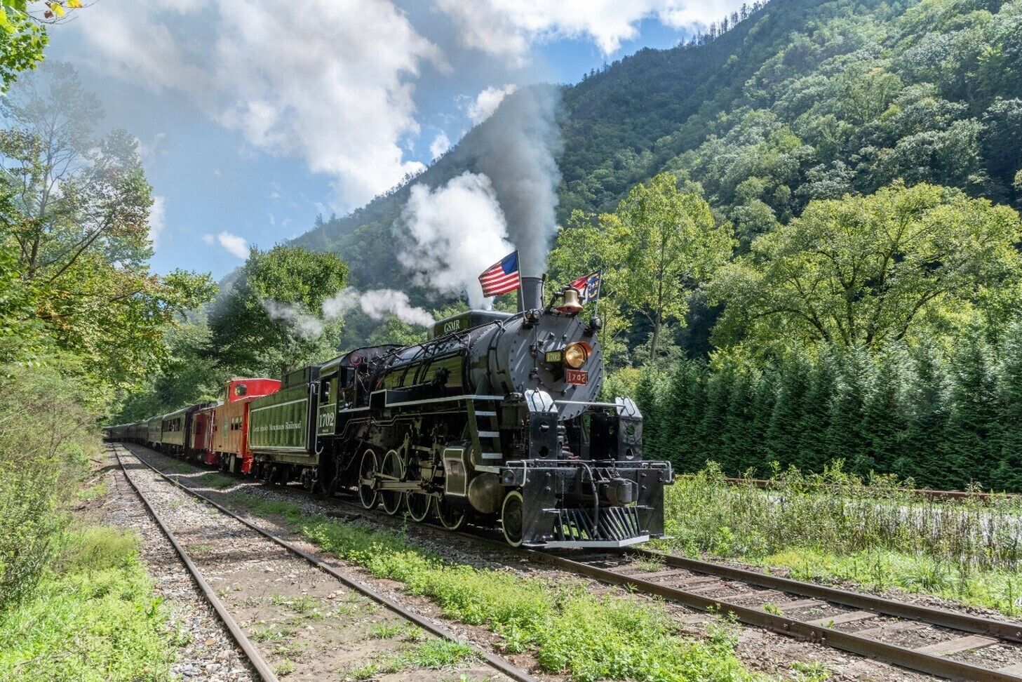 Une locomotive à vapeur, ornée d’un drapeau américain, se trouve sur une voie ferrée, entourée d’une végétation luxuriante et de montagnes.