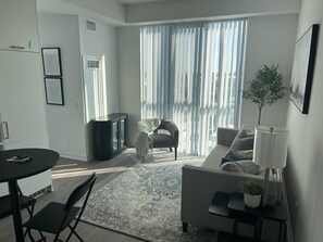 Un salon moderne comprenant un canapĂ©, une table Ă manger, un tĂ©lĂ©viseur et une grande fenĂȘtre Ă©quipĂ©e de stores.