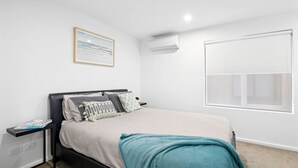 A bedroom with a bed, a bedside table, a wall-mounted air conditioner, a window, and a framed picture on the wall. - Oceania 8 (Lorne)