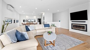A modern living room with a sectional sofa, a flat-screen TV, a fireplace, and a dining area in the background. - Oceania 8 (Lorne)