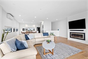 A modern living room with a sectional sofa, a flat-screen TV, a fireplace, and a dining area in the background. - Oceania 8 (Lorne)