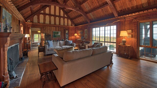 A spacious living room with a high wooden ceiling, a fireplace, and large windows offering a view of the outdoors. - Large Historic Home with views of Crystal Lake & Lake Michigan (Frankfort)