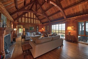 Ein geräumiges Wohnzimmer mit hoher Holzbalkendecke, Kamin und großen Fenstern mit Blick ins Freie.