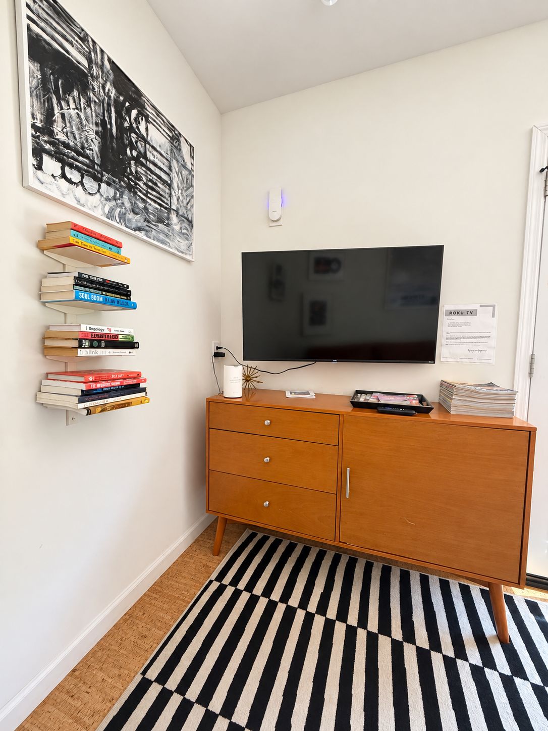 Una habitación con una televisión de pantalla plana montada en la pared, un mueble de madera con cajones, una alfombra a rayas blancas y negras y un estante en la pared con libros.