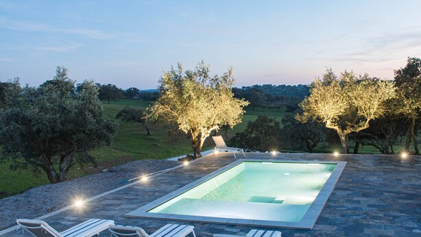 A rectangular pool with two lounge chairs and a table, surrounded by stone tiles and trees.