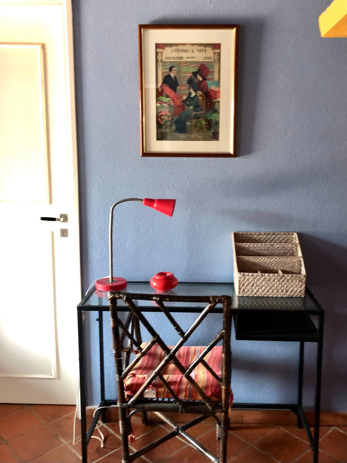 Een kamer met een lijstafbeelding aan de muur, een tafel met een lamp en een rieten mand.