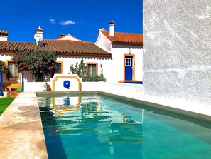 A swimming pool with a clear blue water surface, surrounded by white buildings with orange trim and blue doors. - Small Cottage at Alentejo T1+1 with  pool by Portus Alacer (Portalegre)