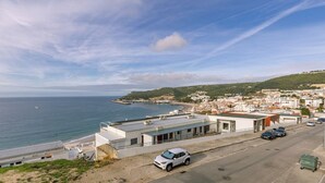 Un poble costaner amb edificis moderns, un carrer amb cotxes aparcats i una vista del mar i les colines al fons.