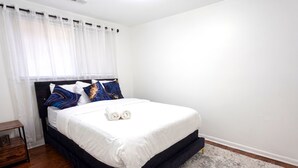 A neatly made bed with white bedding and decorative pillows, a small bedside table, and a window with sheer curtains. - Amazing Mid-Term Rental Entire Home In Glenview (Northbrook)