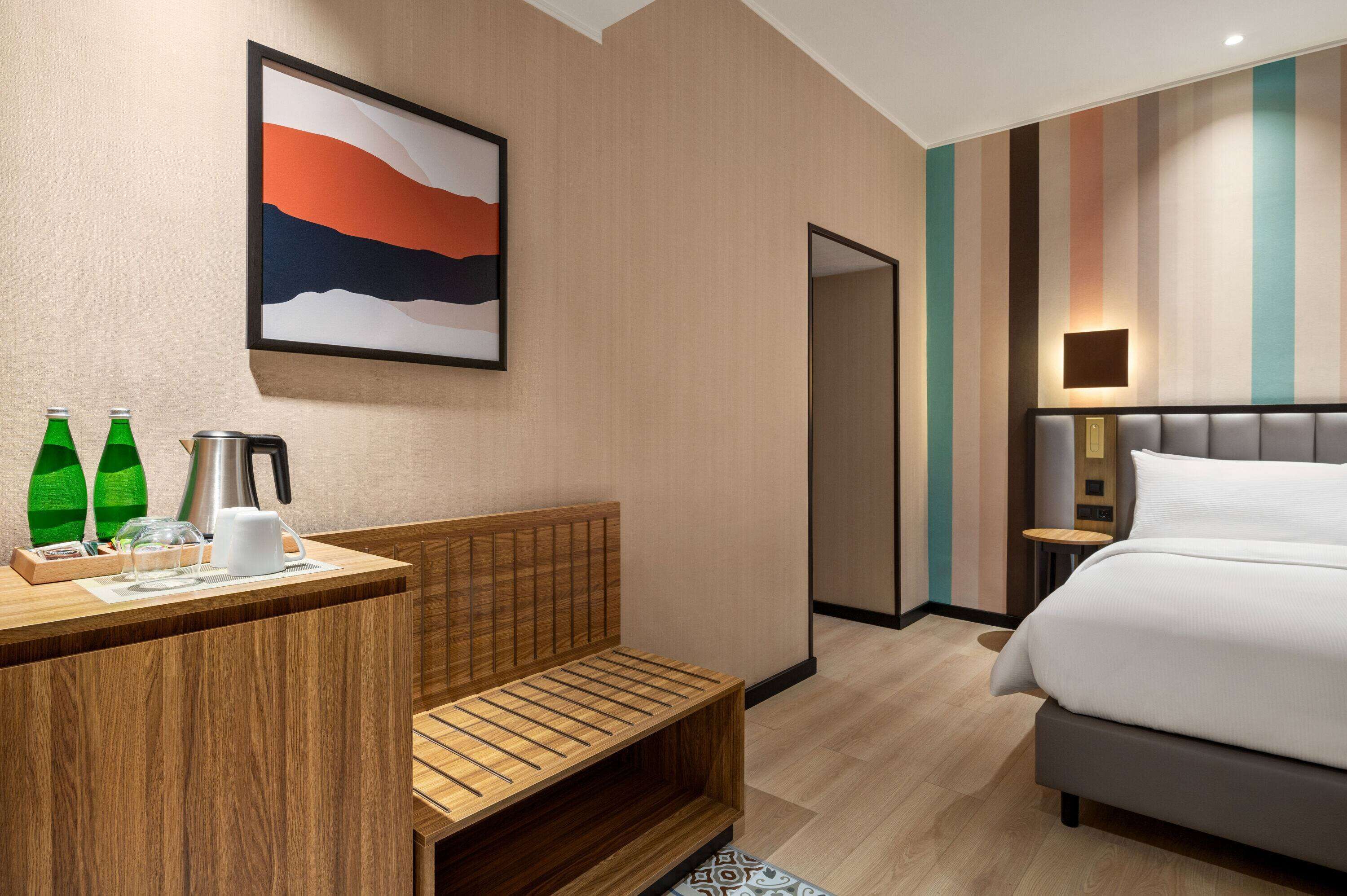 A modern hotel room with a wooden desk, a bench, a bed, and a painting on the wall.