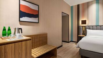 A modern hotel room with a wooden desk, a bench, a bed, and a painting on the wall.