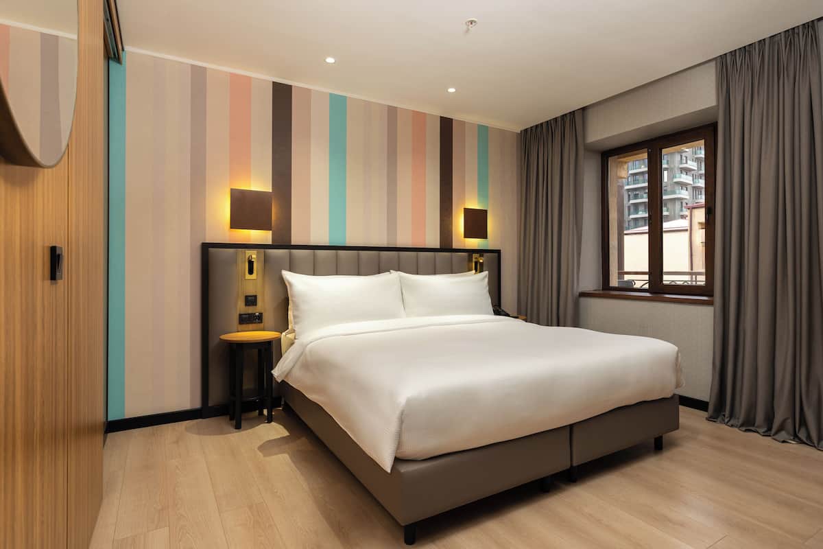 A modern hotel room with a large bed, a nightstand, a wall-mounted lamp, and a window with curtains.
