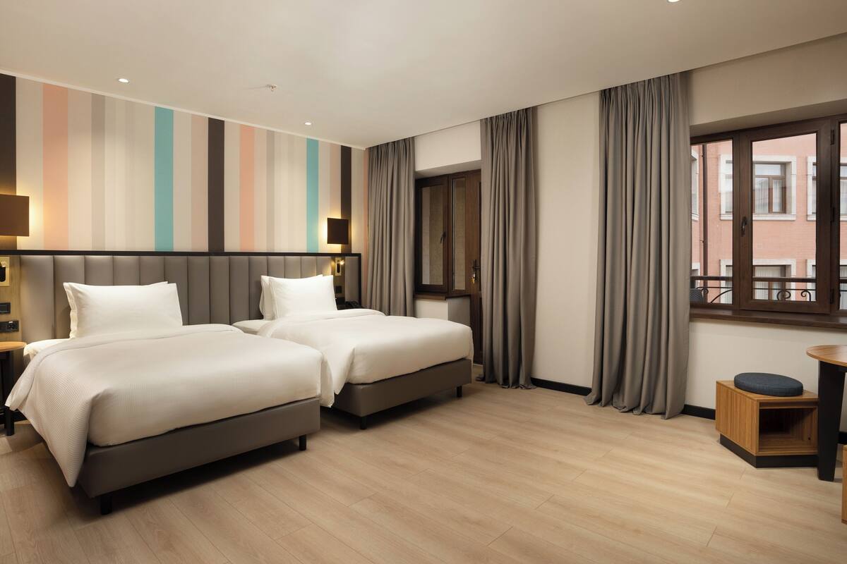 A hotel room with two beds, a small table, a chair, a window with curtains, and a striped wall.