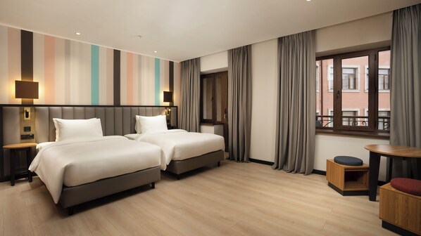 A hotel room with two beds, a small table, a chair, a window with curtains, and a striped wall.