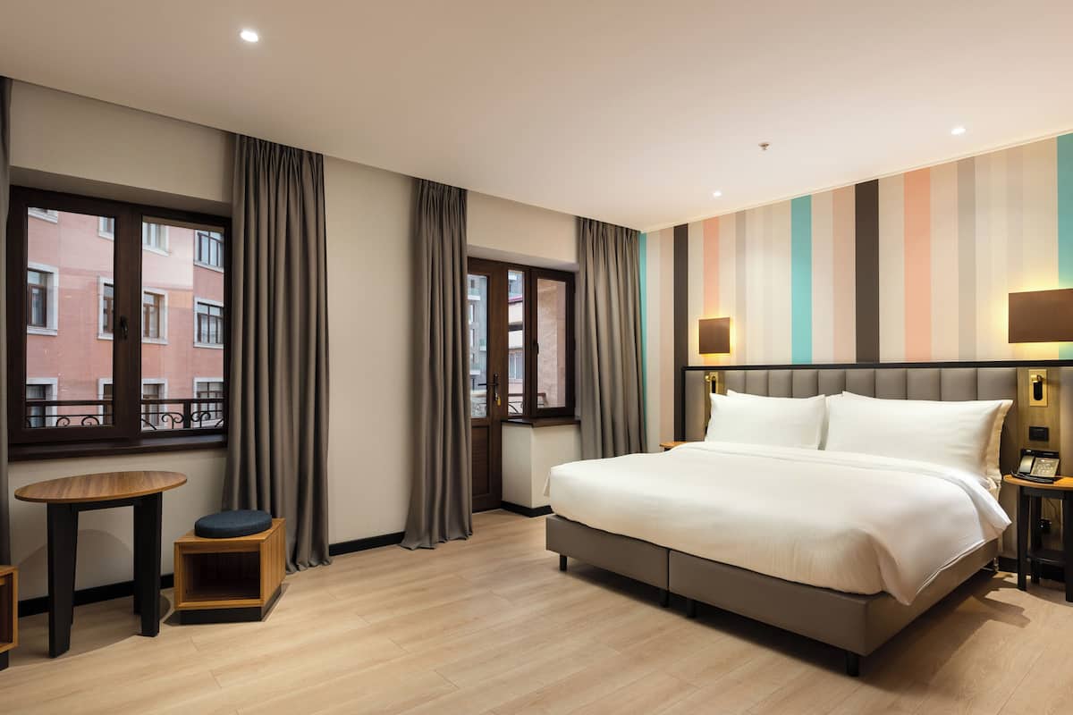 A hotel room with a large bed, a small table, a chair, and a window with curtains.