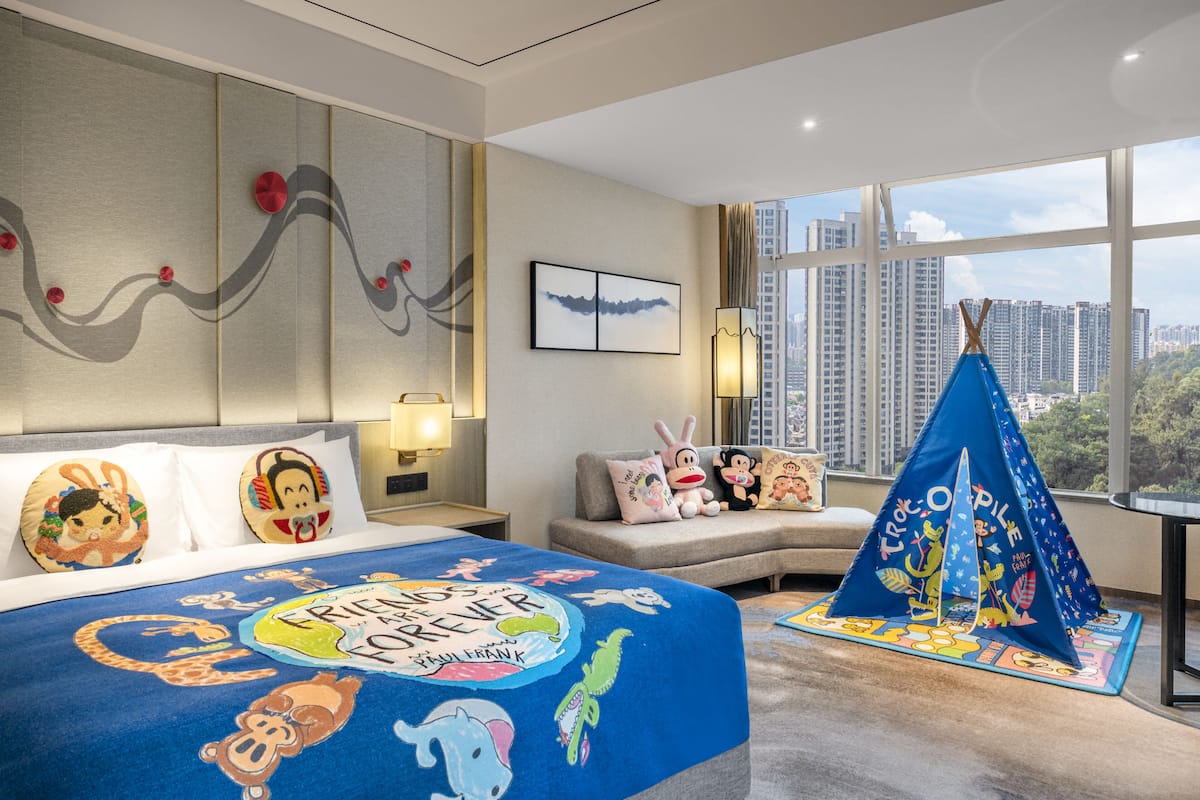 Una habitación de hotel con una cama grande, una manta azul con personajes de dibujos animados, un pequeño sofá, una ventana con vista a la ciudad y una decoración tipi.