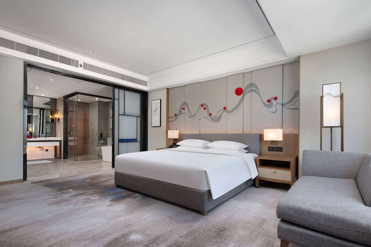 Una habitación de hotel moderna con una cama grande, mesitas de noche, una silla y un baño visible a través de una puerta abierta.