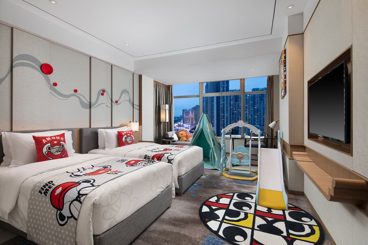 Habitación de hotel moderna con dos camas, un televisor grande, vistas a la ciudad y una zona de juegos para niños.