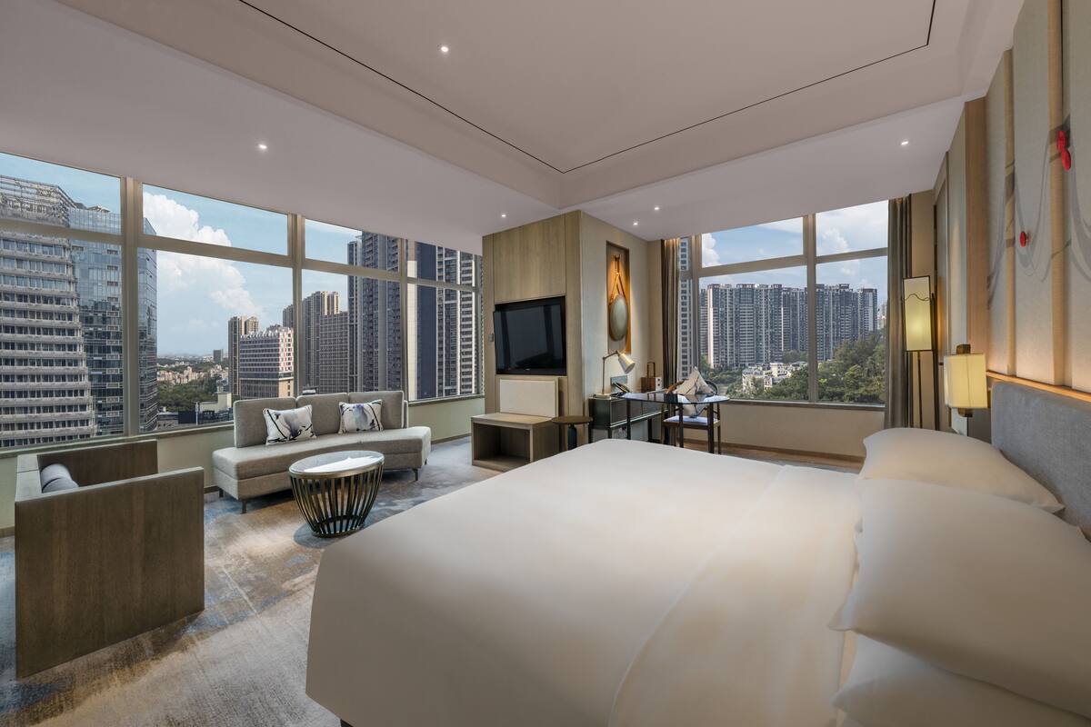 Una habitación de hotel moderna con una cama grande, un sofá, un escritorio y un televisor. La habitación tiene ventanas amplias con vistas a la ciudad.