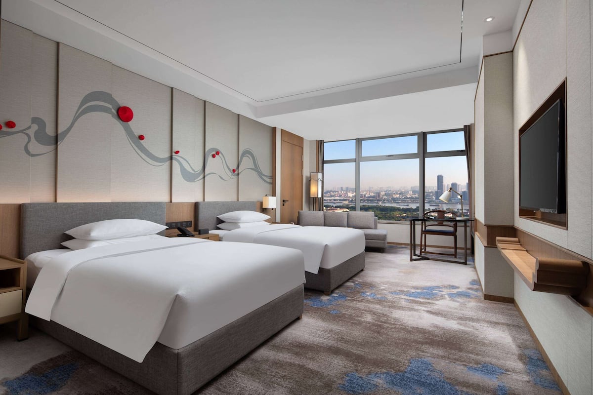 Habitación de hotel con dos camas, un sofá, un televisor y un amplio ventanal con vistas a la ciudad.