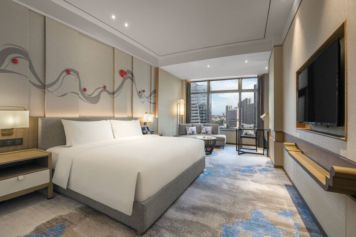 Una habitación de hotel moderna con una cama grande, un escritorio, una silla, un televisor y vistas a la ciudad.