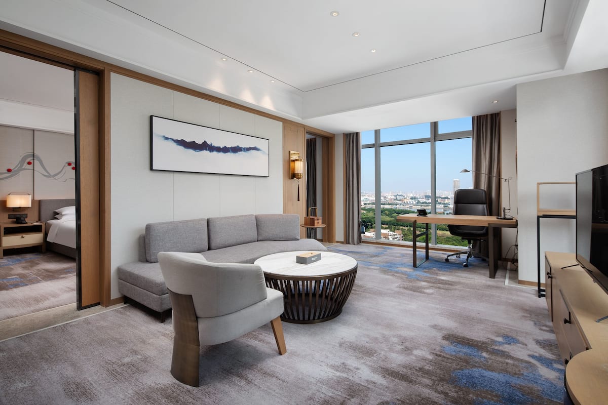 Una habitación de hotel moderna con un ventanal grande, un sofá, una mesa de centro redonda, un escritorio con una silla y un televisor.