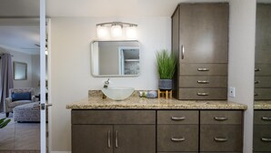 Un baño con encimera de granito, lavabo sobre encimera, espejo, mueble con cajones y una planta en maceta.