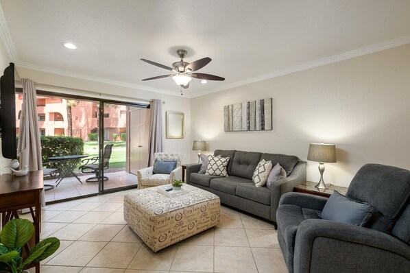 A living room with a grey sofa, a patterned ottoman, a ceiling fan, and a view through a glass door to an outdoor area.