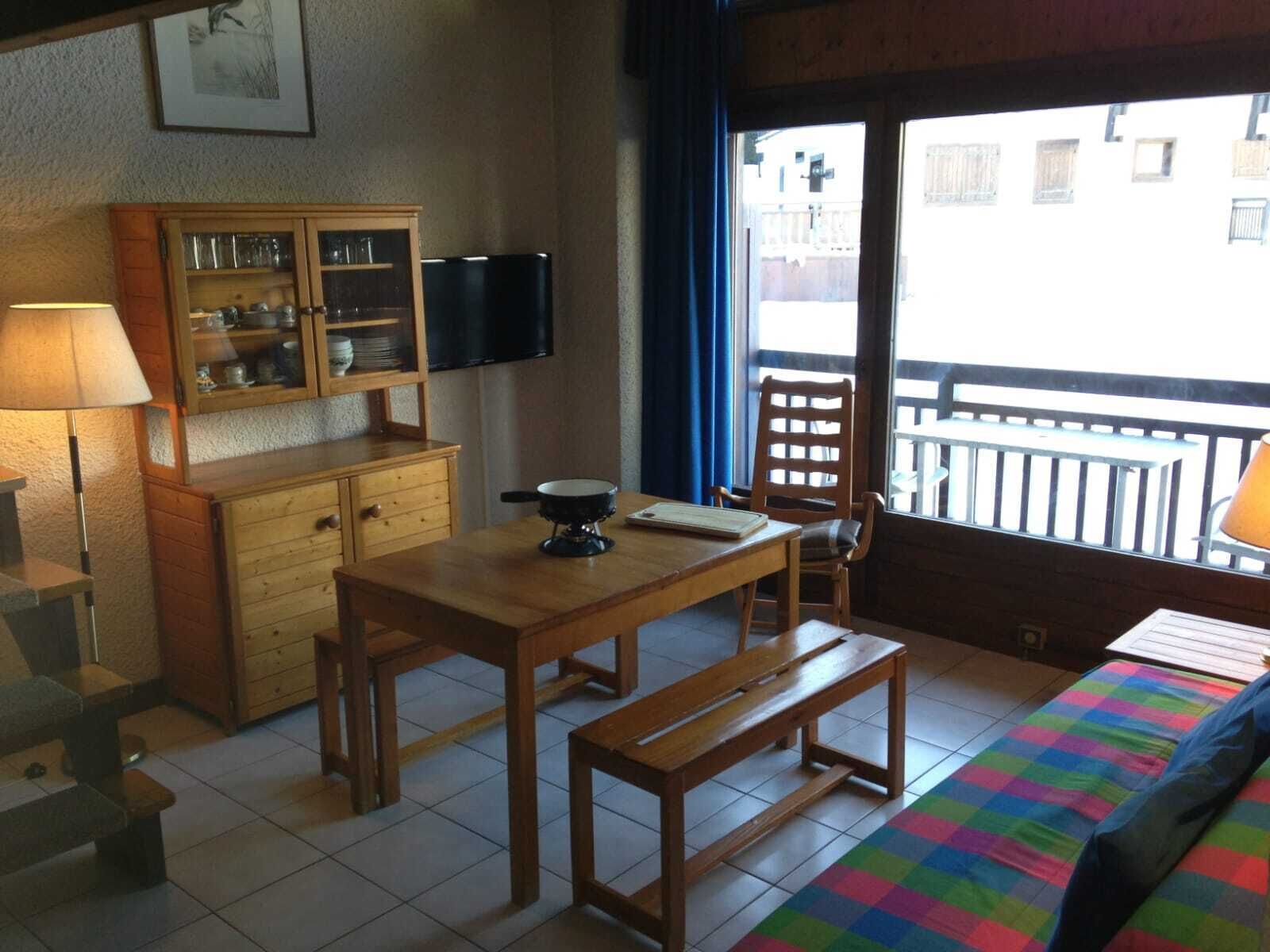 Une pièce chaleureuse avec une table en bois, un banc, un lit recouvert d’une couverture colorée, une armoire aux portes en verre et un téléviseur fixé au mur.