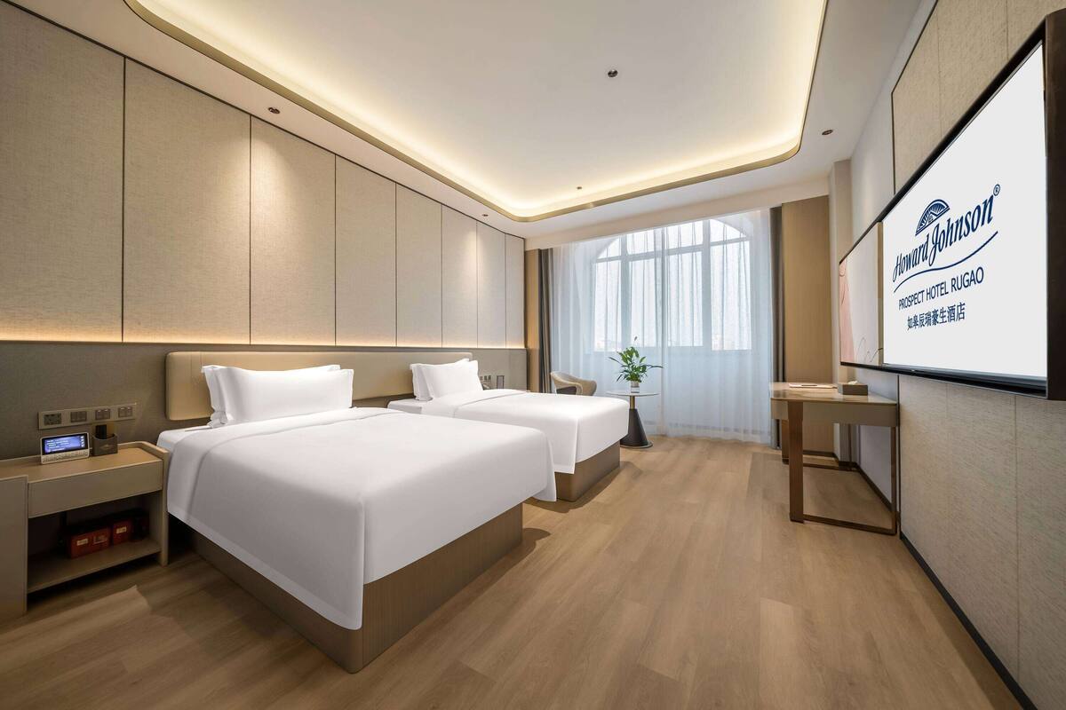 Habitación de hotel con dos camas, un escritorio, una silla, una planta y una pantalla grande que muestra el nombre del hotel.