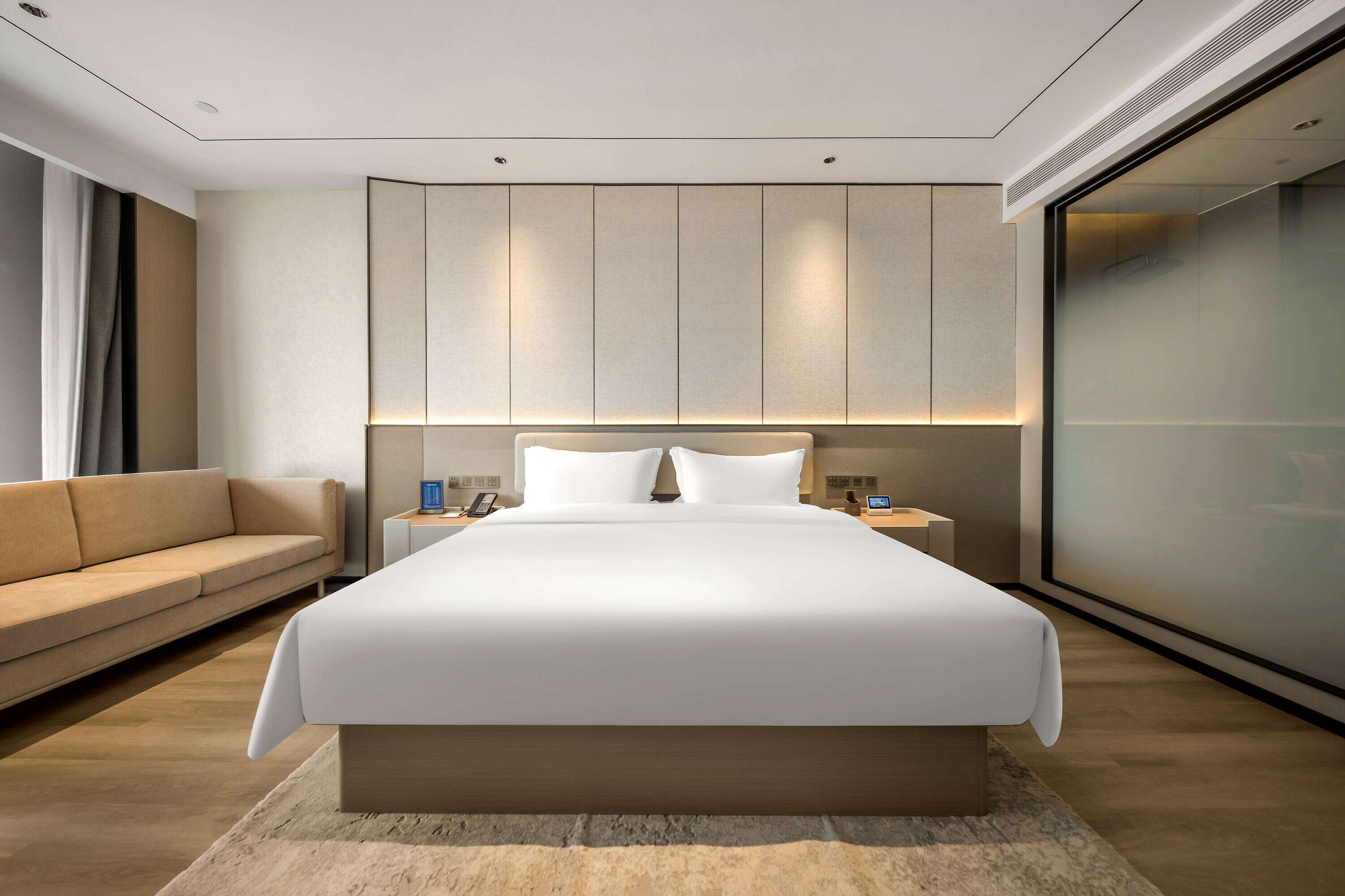 Una habitación de hotel moderna con una cama grande, un sofá, una mesita de noche y un ventanal amplio.