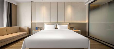 Una habitación de hotel moderna con una cama grande, un sofá, una mesita de noche y un ventanal amplio.