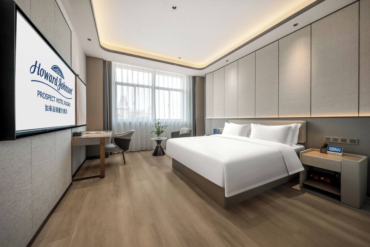 Una habitación de hotel moderna con una cama grande, un escritorio y una pantalla de televisión mostrando el nombre del hotel.