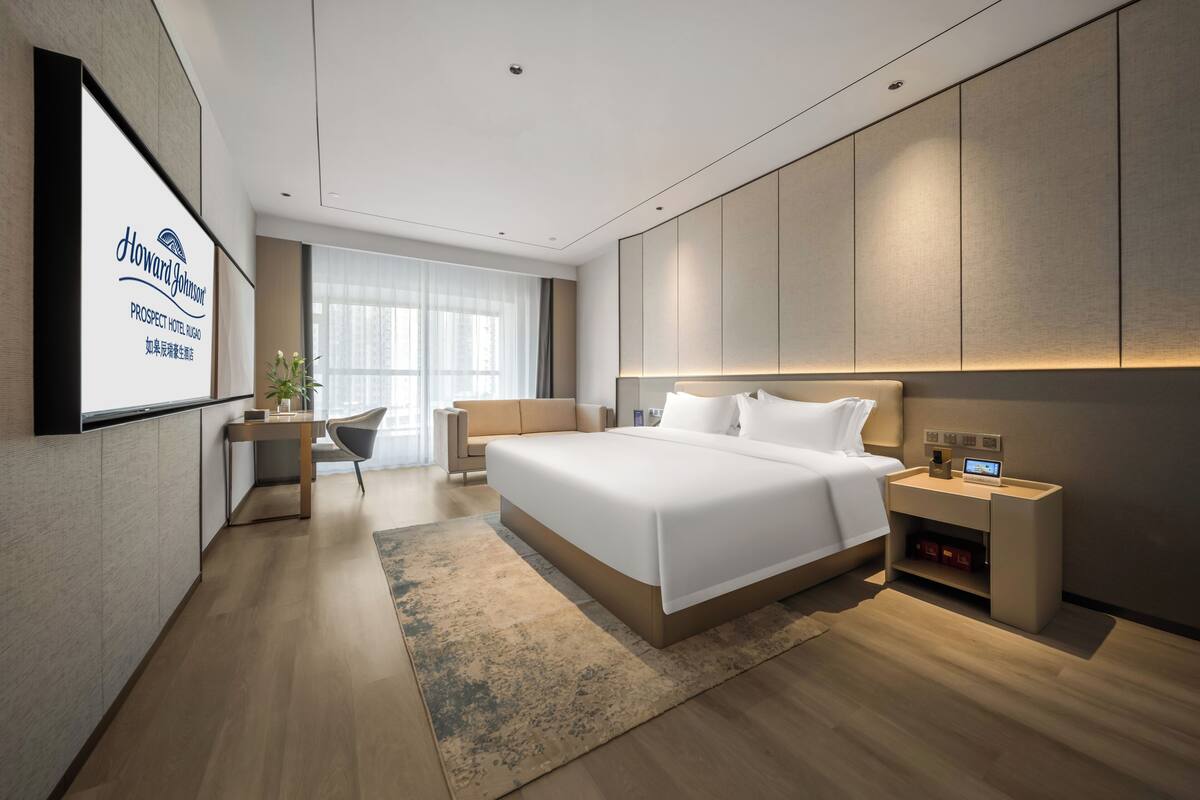 Una habitación de hotel moderna con una cama grande, mesitas de noche, un escritorio y un amplio ventanal con cortinas.