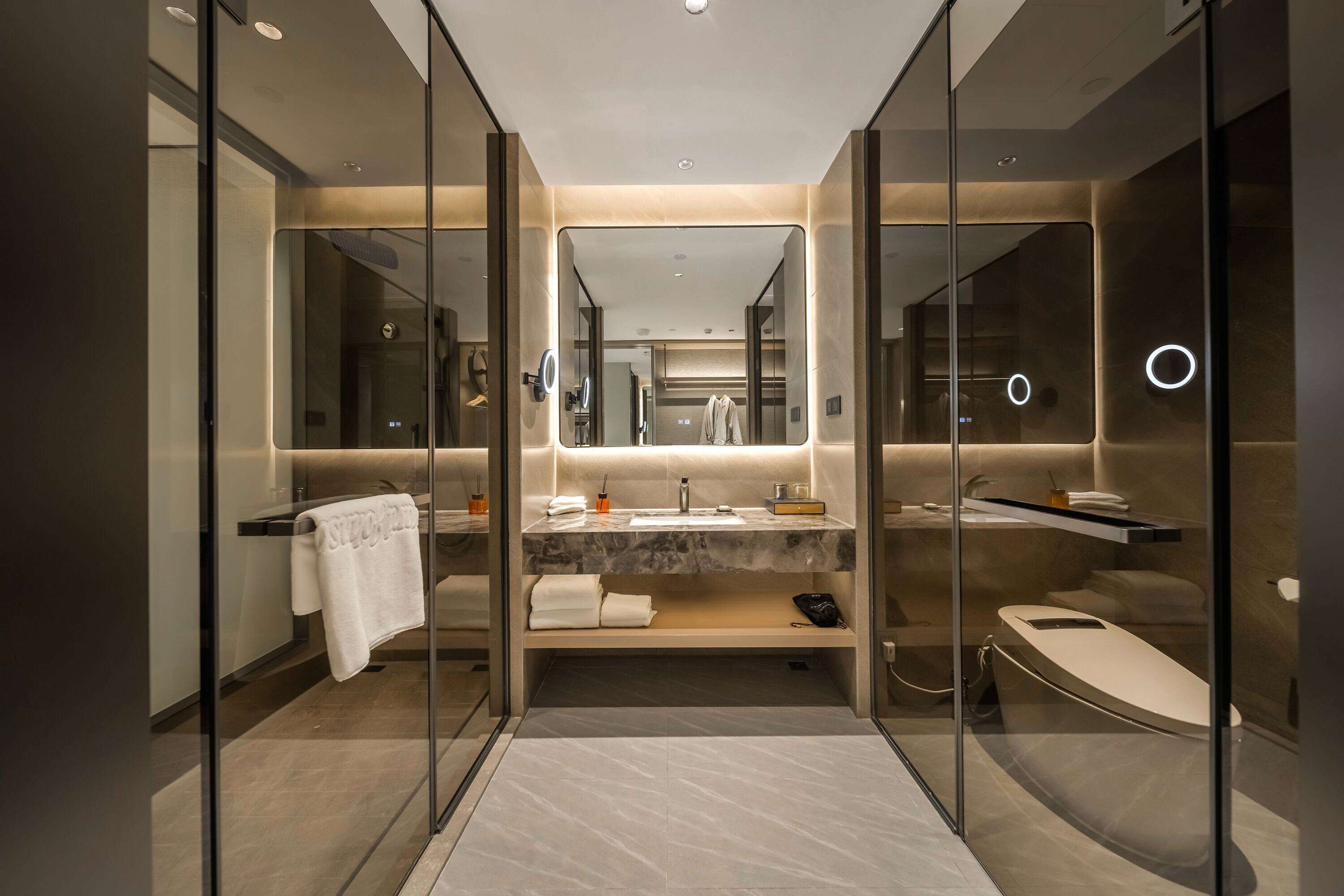 A modern bathroom with a glass partition, a vanity with a marble countertop, a bathtub, and a large mirror.