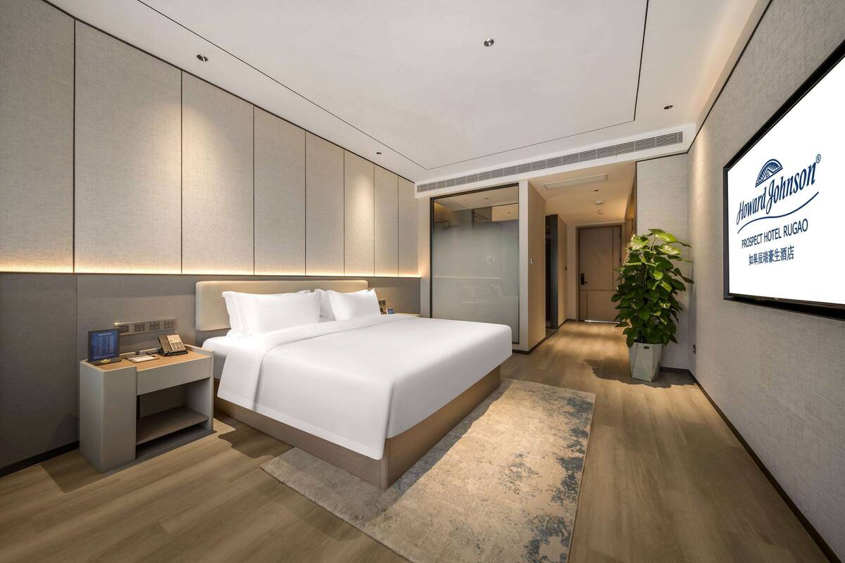 Habitación de hotel moderna con una cama grande, una mesita de noche con teléfono, una mesita auxiliar, una planta en maceta y una pantalla mural que muestra el nombre del hotel.
