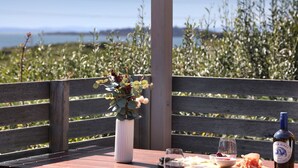 A table with a bottle of wine, glasses, and a bowl of fruit on a wooden bench with a view of a field and a body of water. - Bayview Bliss in Surf Beach (Surf Beach)
