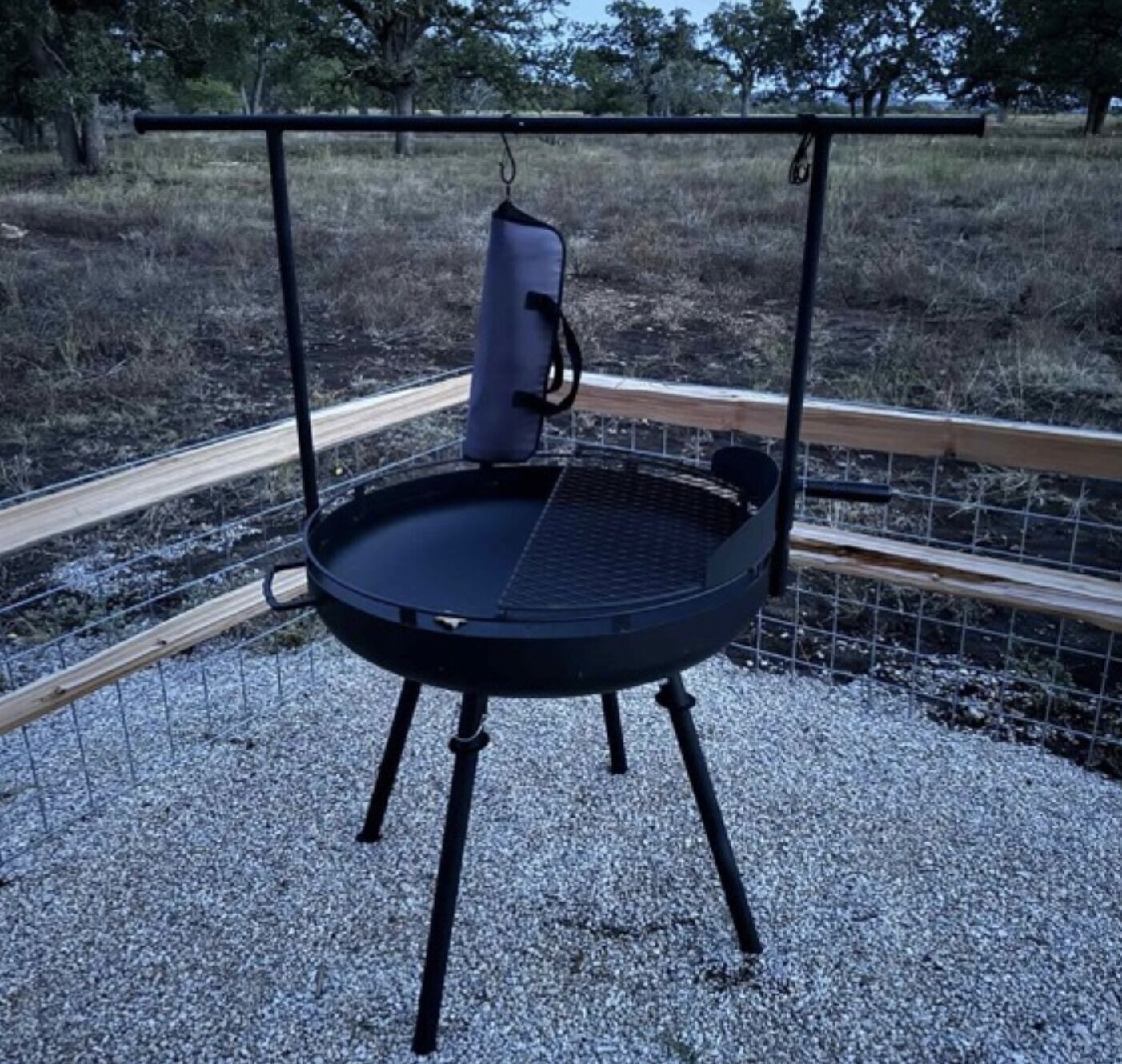 Un barbecue portable doté d’une grille en métal et d’un support à casseroles suspendu, installé sur une surface de gravier, avec une clôture en bois en arrière-plan.