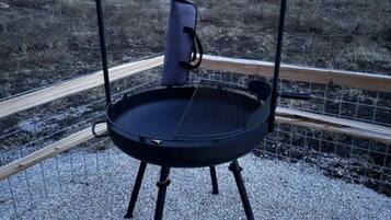 Un barbecue portable doté d’une grille en métal et d’un support à casseroles suspendu, installé sur une surface de gravier, avec une clôture en bois en arrière-plan.