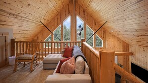 Une charmante cabane en bois avec une chambre à l’étage, dotée d’une grande fenêtre, d’un fauteuil à bascule, d’un canapé et d’une cheminée.