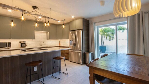 A modern kitchen with a central island, bar stools, a dining table, and a large window with a view of the outdoors. - NEW! Stylish & Cozy Sea Grape near Beach & Flagler (New Smyrna Beach)