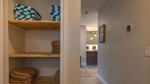 A hallway leading to a kitchen with wooden shelves holding folded towels and a checkered blanket. - NEW! Stylish & Cozy Sea Grape near Beach & Flagler (New Smyrna Beach)