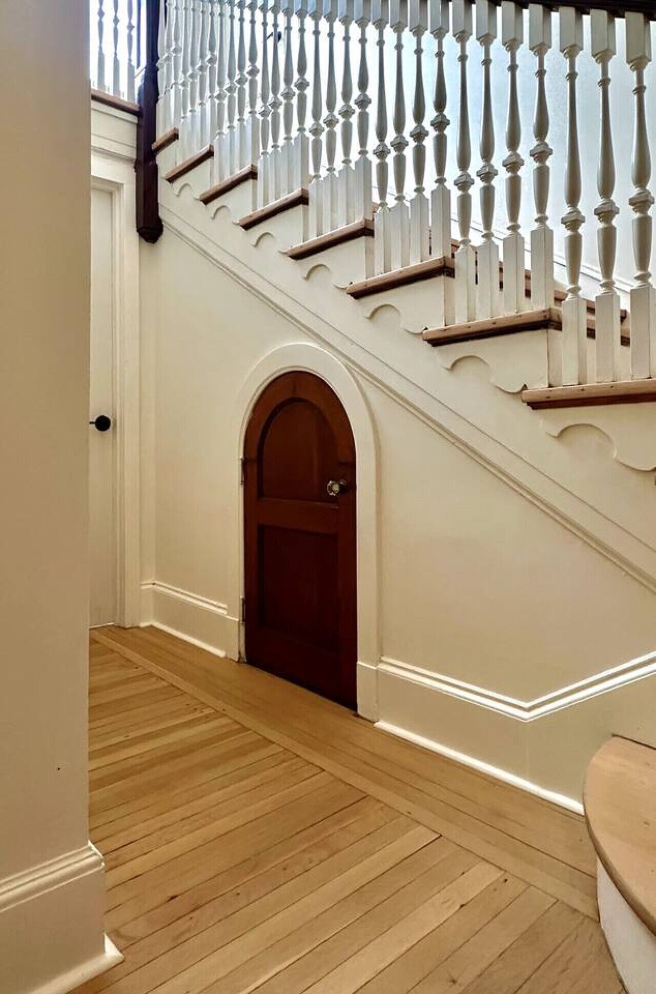 A hallway with a wooden floor leading to a staircase with white balustrades and a wooden door at the bottom.